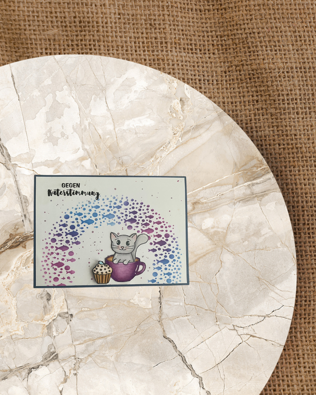 Karte mit einer Katze in einer Tasse sitzend. Daneben ein Cupcake mit einem Fisch. Im Hintergrund ein Halbkreis Fische mit Wasserblasen in Blau/Lila. Spruch gegen Katerstimmung.