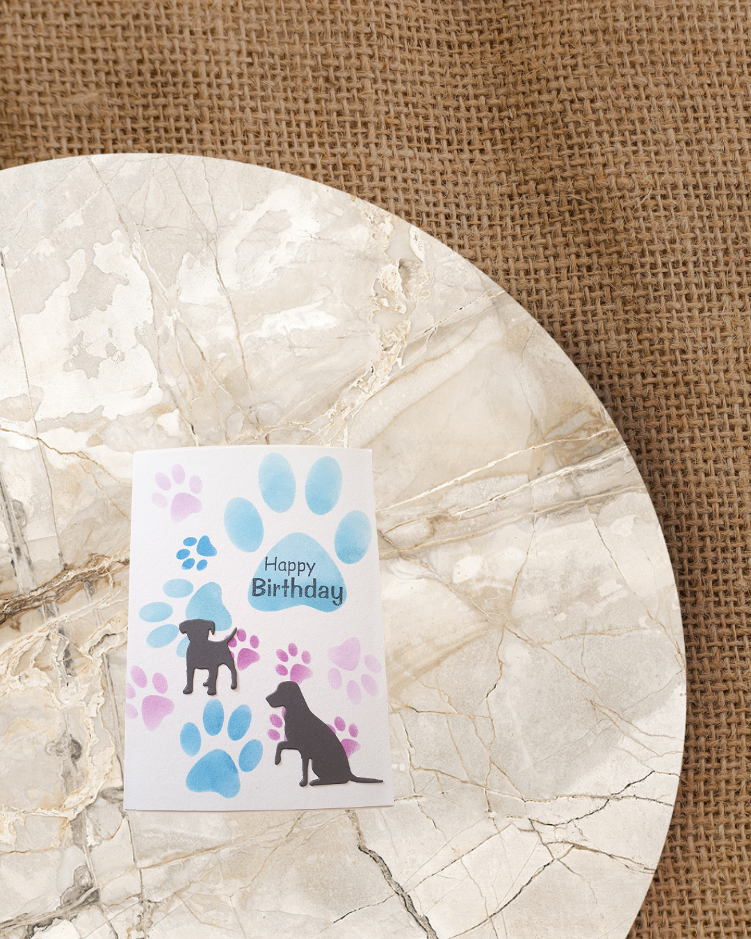 Geburtstagskarte mit bau und lila Pfoten abdrücke in verschiedenen Größen im Hintergrund. Ein stehender und sitzender Hund als schwarze Silhouette und Happy Birthday.