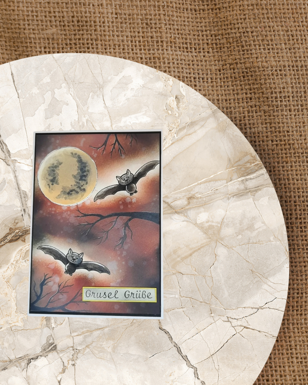 Zwei liegende Fledermäuse auf einer Karte zu Halloween. Im Hintergrund 3 Äste und ein Mond. Die Farben sind in Braun, Schwarz und Orange gehalten. Spruch Grusel Grüße