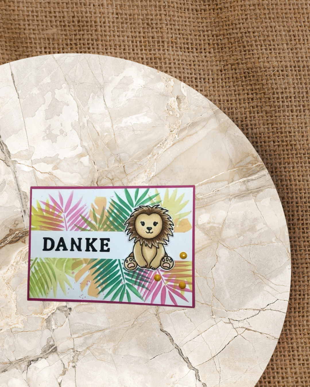 Löwe sitzend rechts in der Mitte. Ein weißer Steifen in der Mitte mit dem Spruch Danke. Im Hintergrund in verschiedenen Farben Tropische Blätter