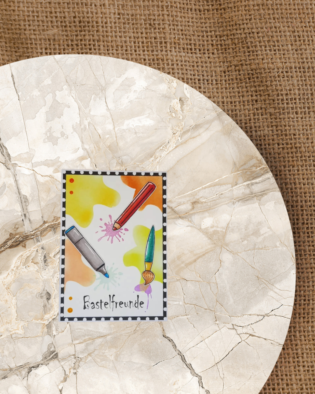 Auf einer Karte verschiedene Stifte und ein Pinsel im Hintergrund verschiedenfarbige Kleckse. Als Spruch Bastelfreunde