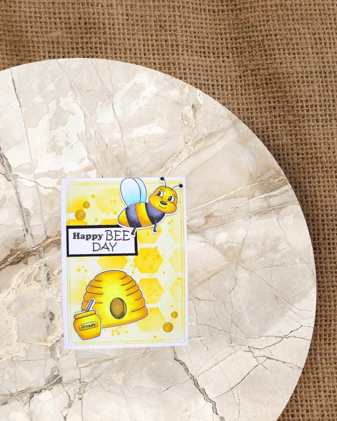 Karte mit fliegender Biene dazu ein Bienenstock und Honigtopf. Im Hintergrund gelbe Waben. Spruch Happy Bee Day