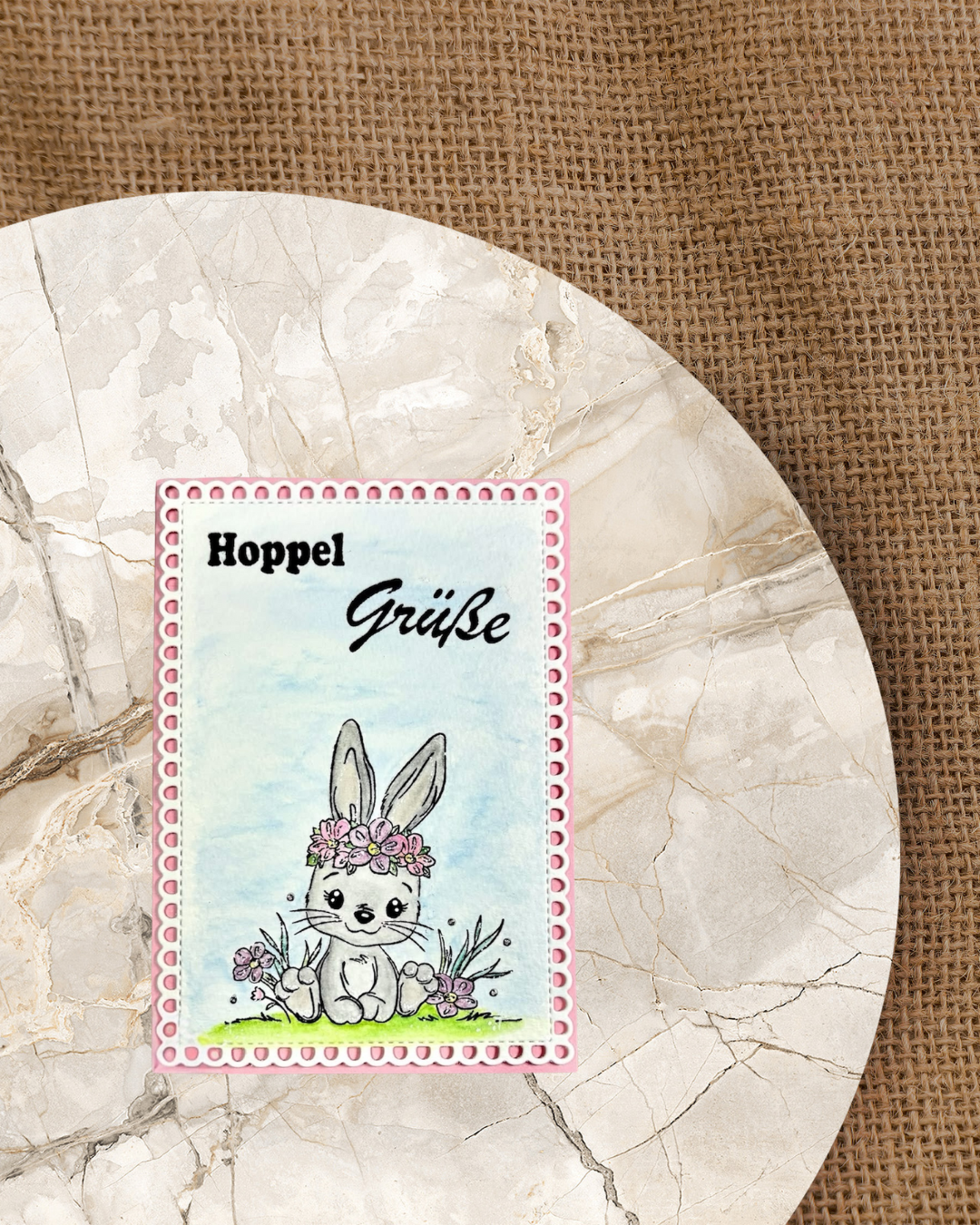 Sitzendes Hasenmädchen mit Blumenkranz auf Graß und dem Spruch Hoppel Grüße