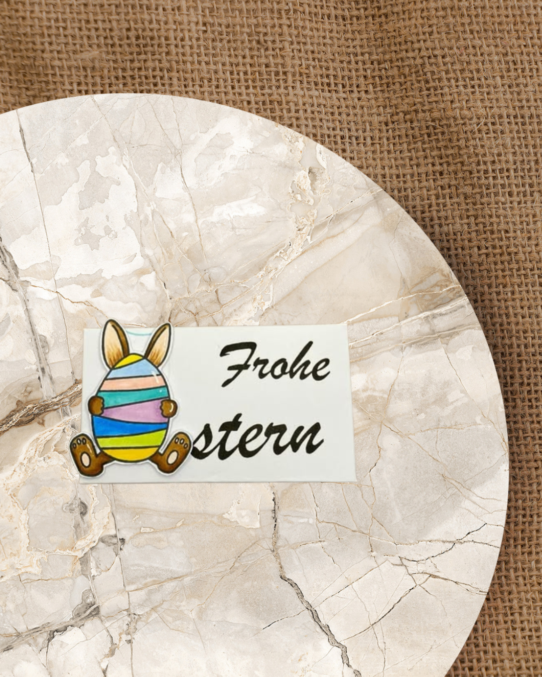 Auf einer Geschenkbox ist ein großer Schriftzug Frohe Ostern. Das O in Ostern ist ein Ei mit Hasenhoren und Füßen. Das Ei ist bunt bemalt