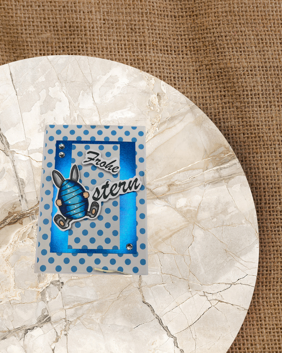 Blaue Karte mit Großem Schriftzug Frohe Ostern. Das O in Ostern ist ein Ei mit Hasenhoren und Füße