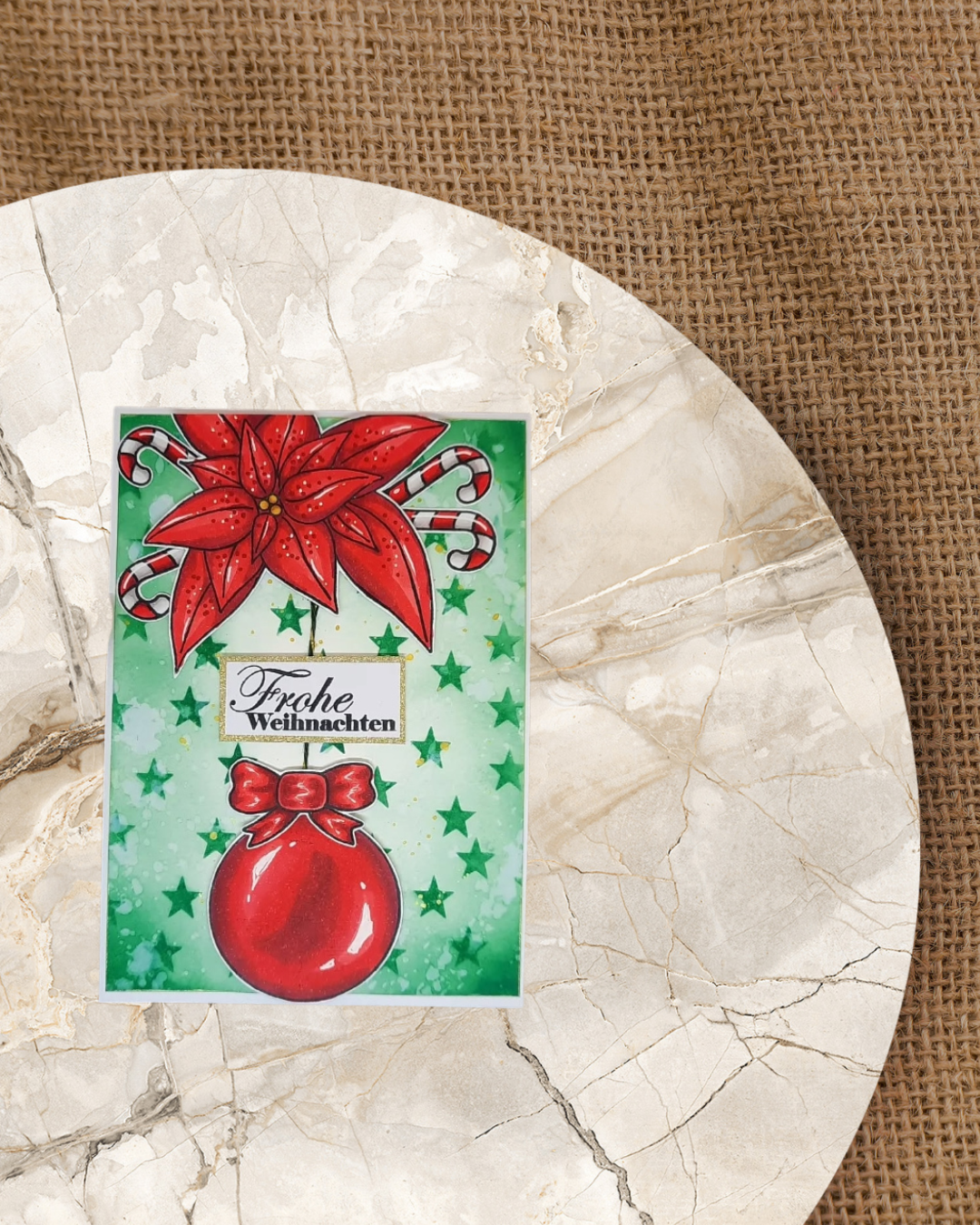 Karte mit einer roten hängenden Weihnachtskugel. Oben ein roter Weihnachtsstern mit Zuckerstangen. Im Hintergrund grüne kleine Sterne. Spruch Frohe Weihnachten