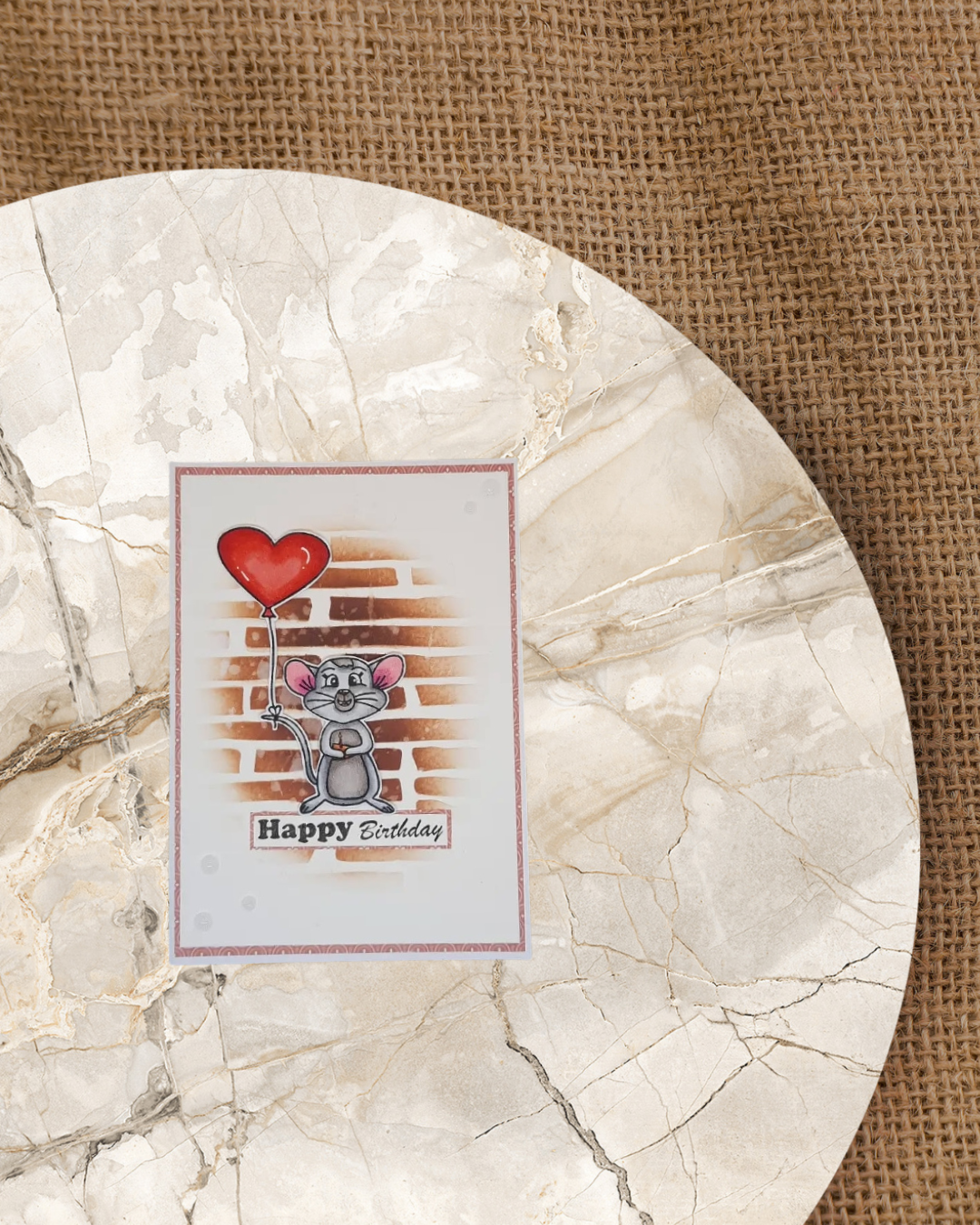 Auf einer Karte eine Maus stehend mit einem Käsestück und Kerze in der Pfote. Am Schwanz ein Luftballon angebunden. Im Hintergrund eine braune Mauer und dem Spruch Happy Birthday