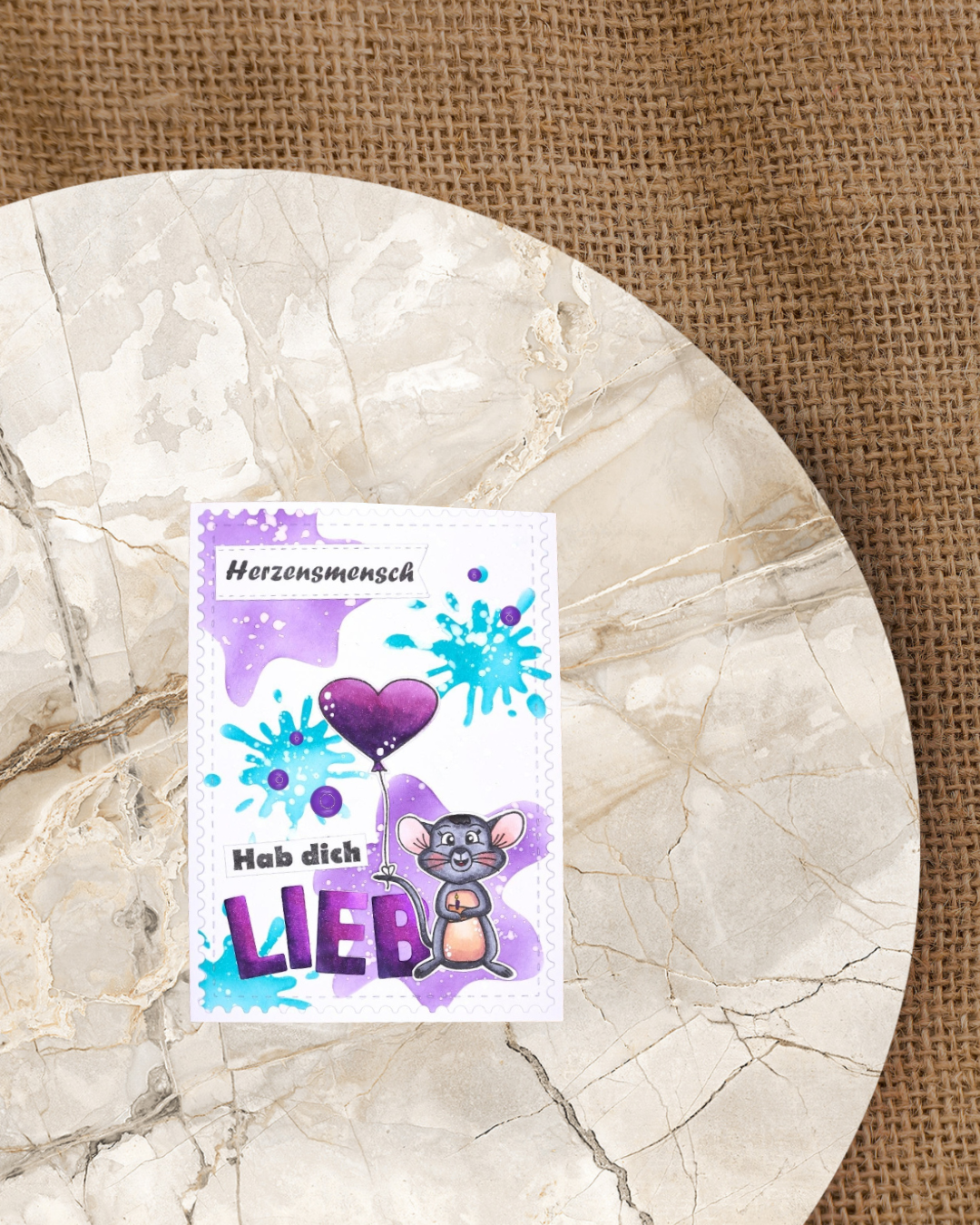 Auf einer Karte eine Maus mit Käsestück und Kerze, am Schwanz ein Luftballon angebunden. Im Hintergrund verschieden Kleckse in Türkis und Lila