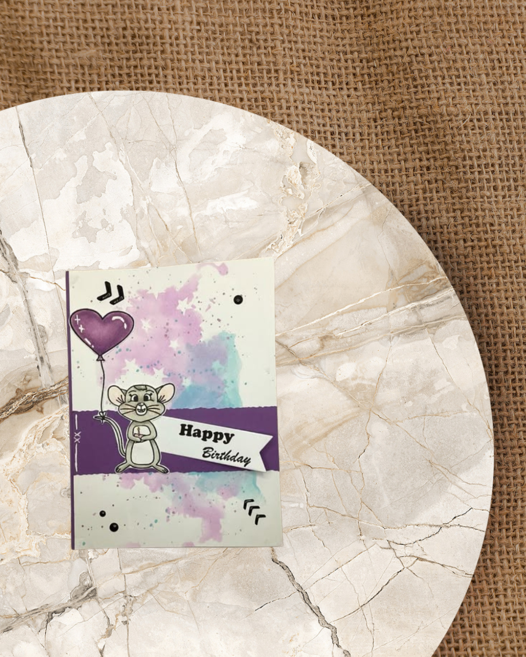 Auf einer Karte eine Maus mit einem Stück Käse mit Kerze. Die Maus hat ein Luftballon am Schwanz. Lila/Blauer Hintergrund mit Sternen und Happy Birthday