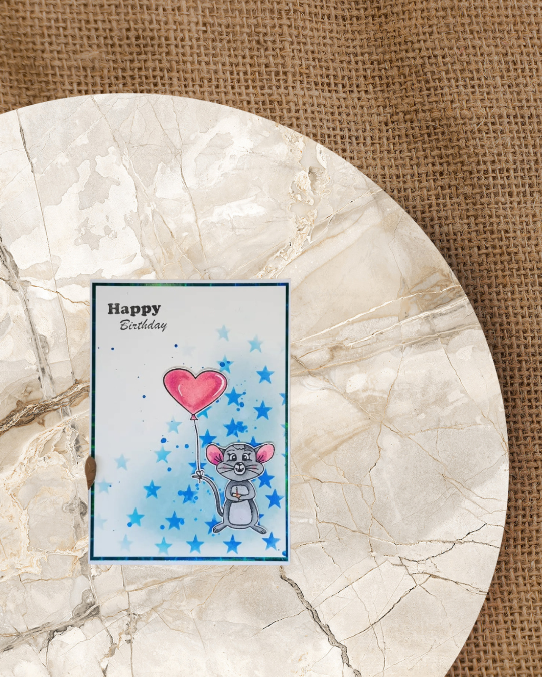 Maus mit Herzballon an den Schwanz gebunden und einem Stück Käse mit Kerze in den Pfoten. Im Hintergrund blaue keine Sterne. Spruch Happy Birthday