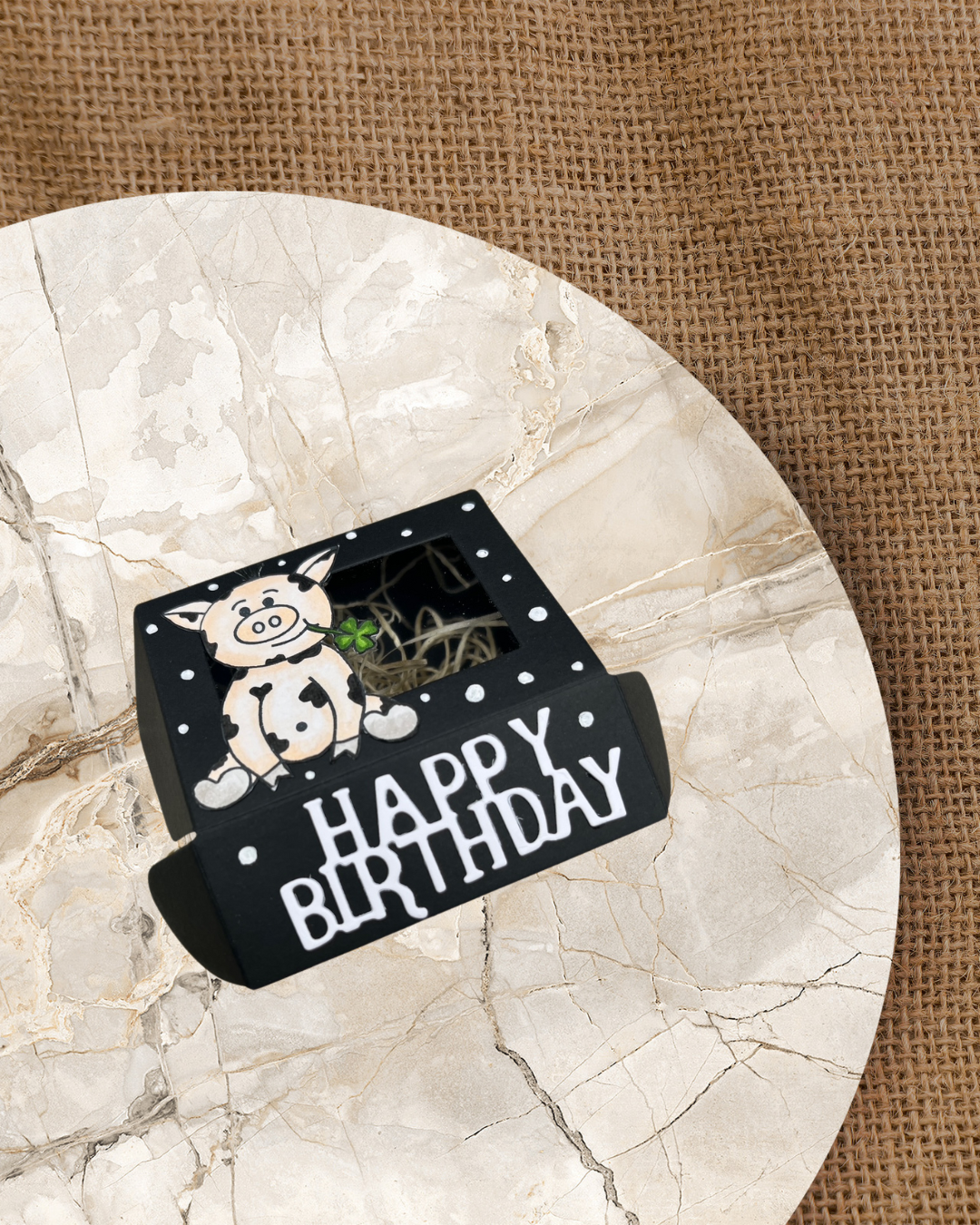 Schwarze kleine Geschenkbox mit weißen Punkten und dem Schriftzug Happy Birthday. Auf der Geschenkbox sitzt ein rosa Schweinchen mit schwarzen Flecken und Kleeblatt aus dem Mund hängend
