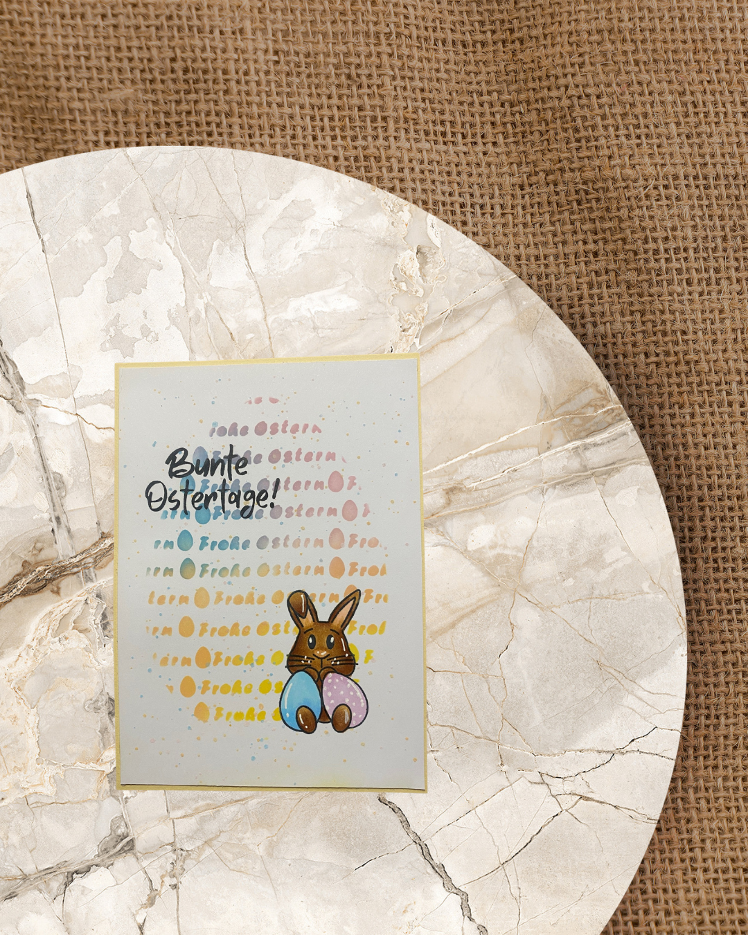 Karte mit einem Hase und zwei Eiern auf dem Schoß. Im Hintergrund bunt Frohe Ostern in Form eines großen Eis und der Spruch Bunte Ostertage