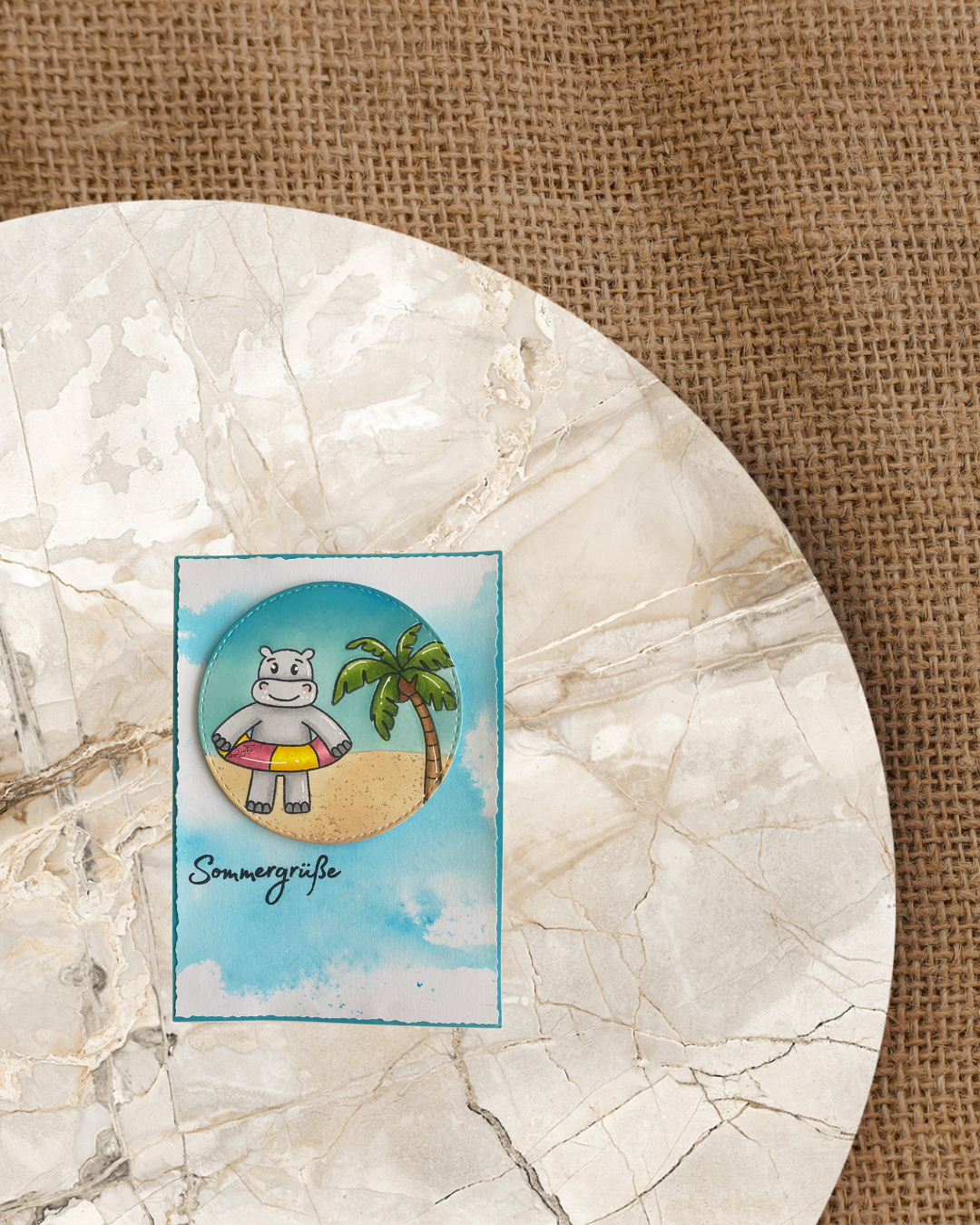 Blauer Schimmer Hintergrund, darauf ein großer ausgestanzter Kreis mit einem Nilpferd mit Schwimmring am Strand mit Palme. Spruch Sommergrüße