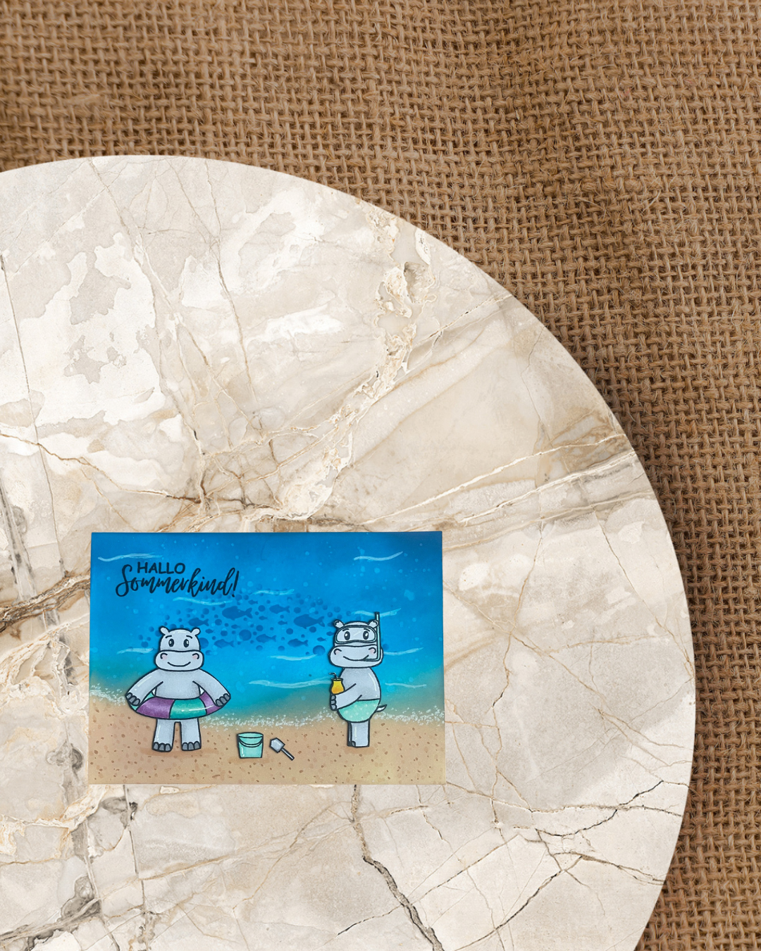 Zwei Nilpferde eins mit Badehose und Cocktail und eins mit Schwimmring Sandeimer und Schaufel am Strand mit Meer. Dunkelblaue Fische und Blubberblasen im Meer. Spruch Hallo Sommerkind