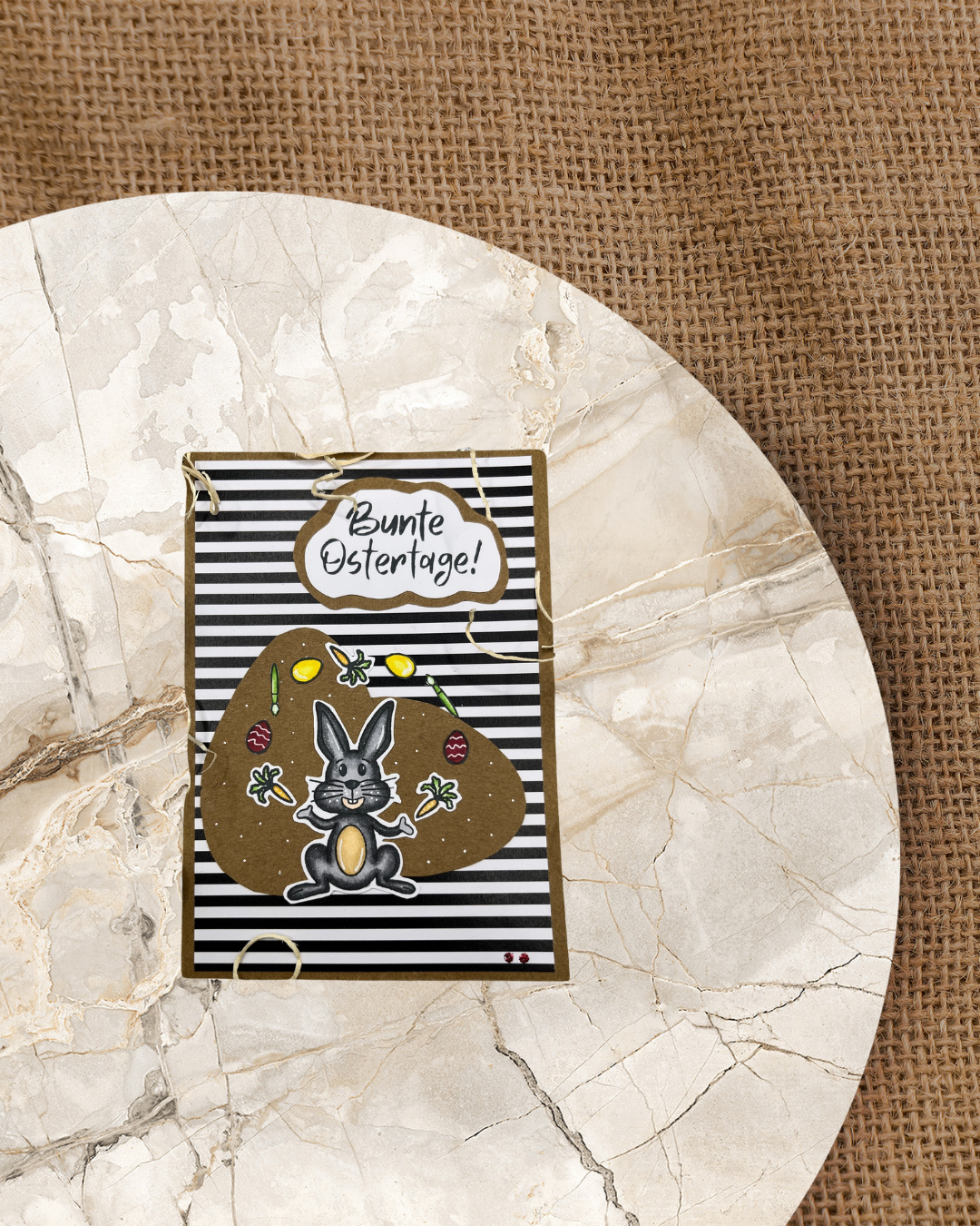 Sitzender jonglierender Hase in grau der Karotten, Eier und Pinsel jongliert. Dazu der Spruch Bunte Ostertage. Der Hintergrund ist schwarz/weiß gestreift