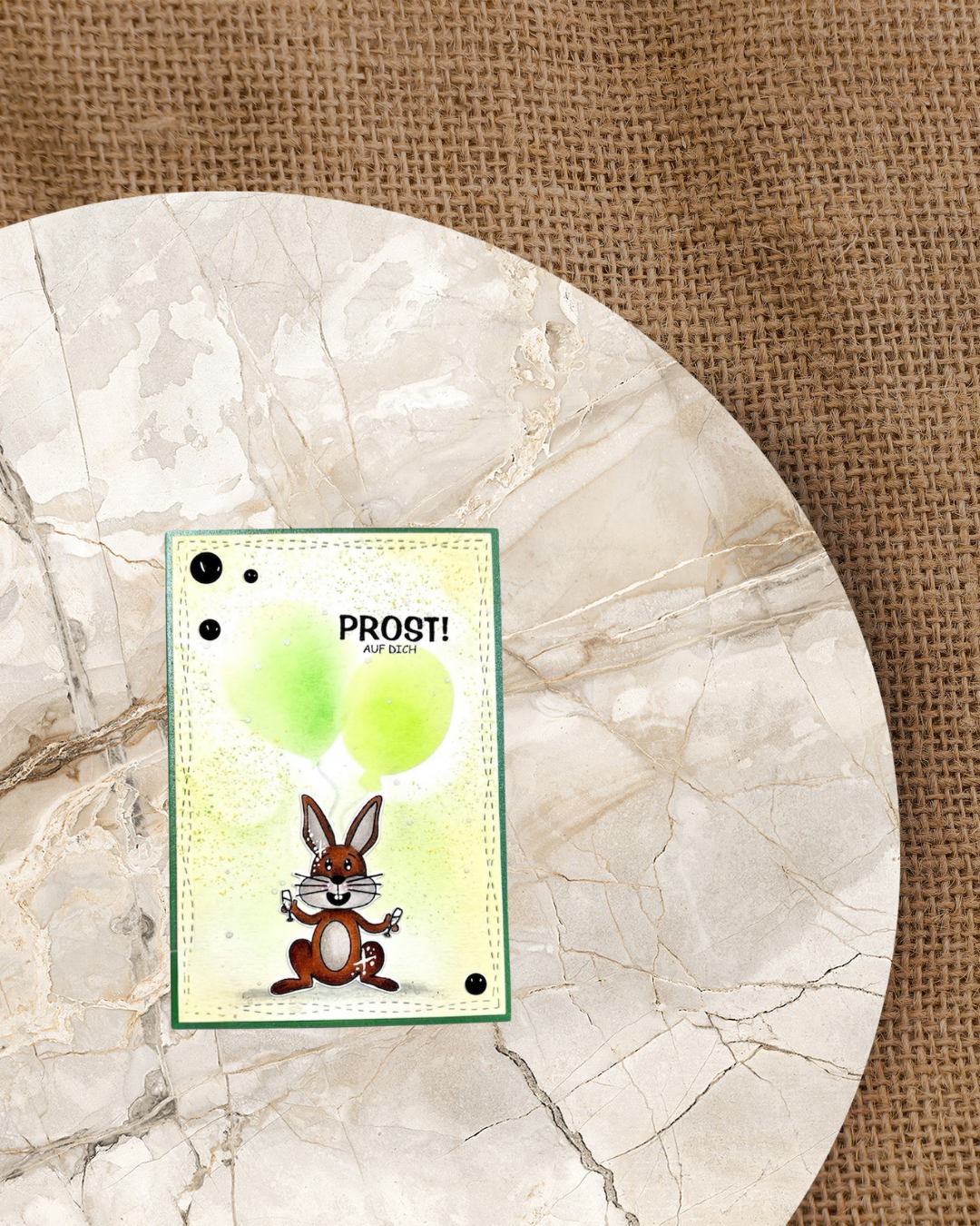 Hase der zwei Sektgläser in den Pfoten hat mit Luftballons im Hintergrund und den Spruch Prost auch dich