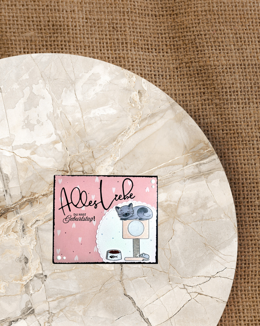 Karte mit rosa mit weißen Herzen Hintergrund. Rechts unten eine weiße Rosette mit einer schlafenden Kate auf einem Kratzbaum mit Fressnapf und Spielmaus nebendran.