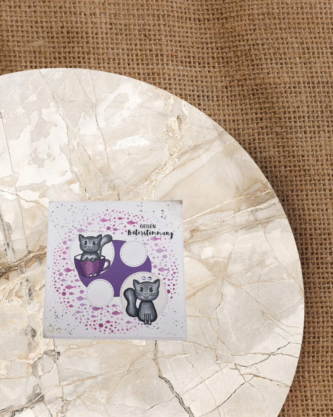 Zwei sitzende Katzen eine davon in einer Tasse. auf einem lila Kreis. Im Hintergrund ein Großer Kreis mit unterschiedlich großen Fischen und Wasserblasen. Als Spruch Gegen Katerstimmung