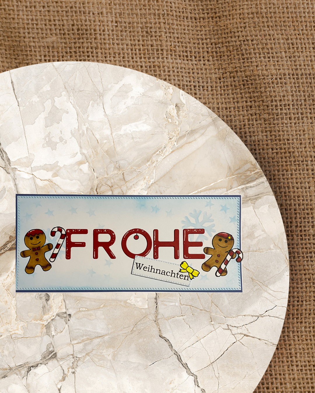 Großer roter Schriftzug Frohe und schräg unterhalb ein Schild mit Weihnachten, links davon ein Lebkuchenmann und rechts eine Lebkuchenfrau mit Zuckerstangen