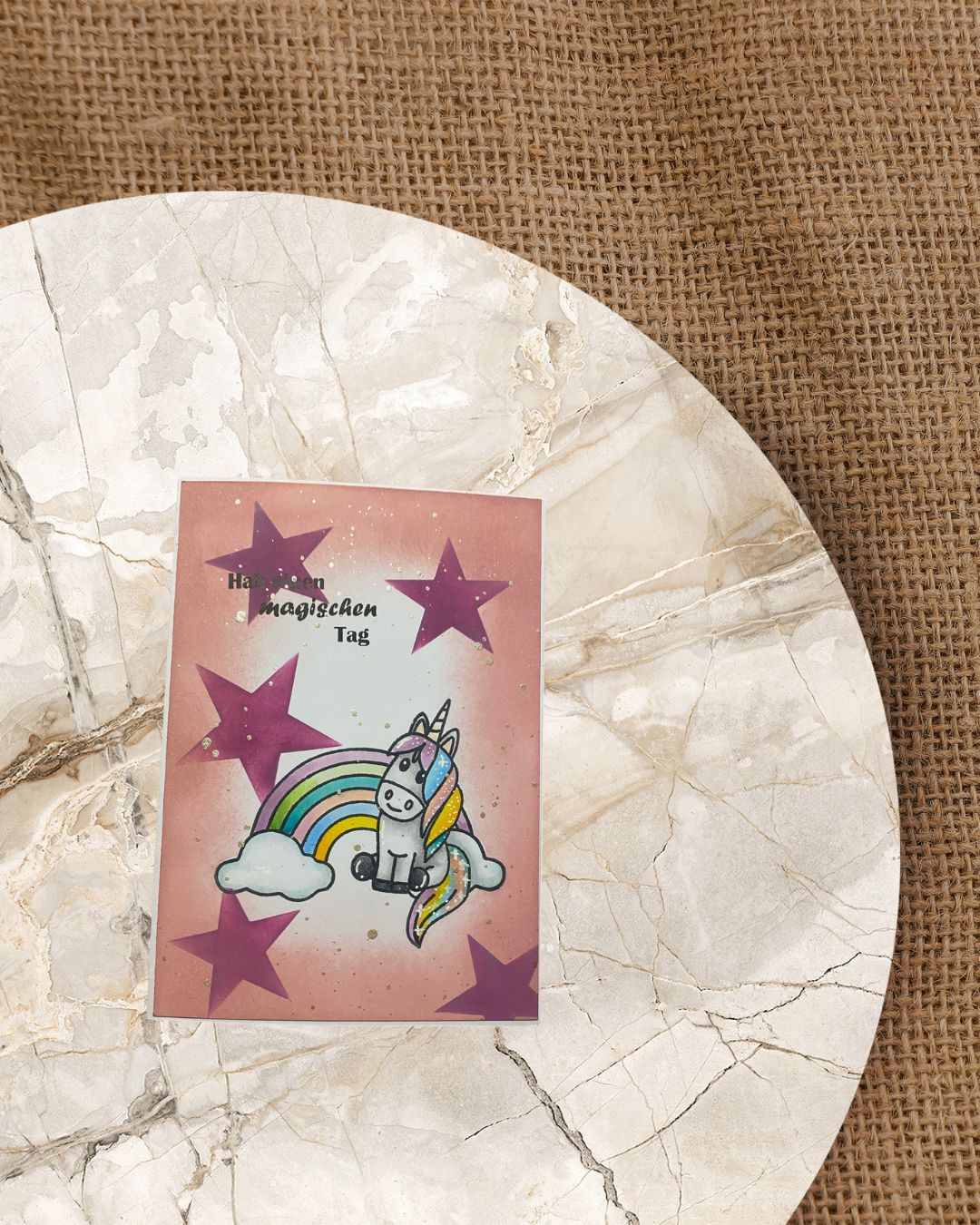 Karte mit lila großen Sternen mit einem sitzenden Einhorn vor einem Regelbogen