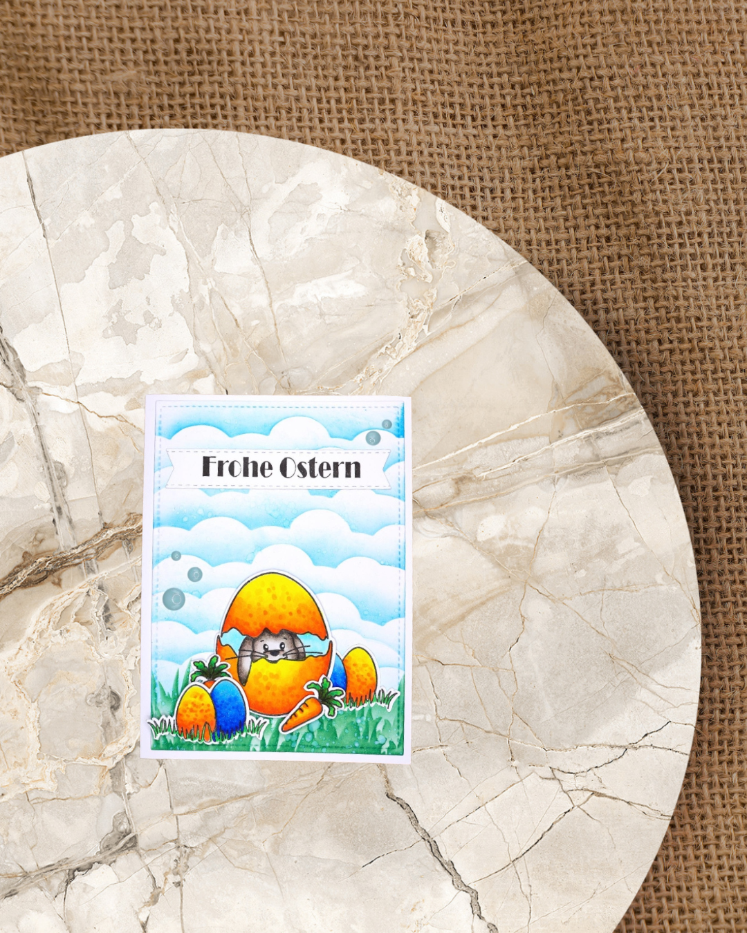 Karte mit einem Hasen der aus einem zerbrochenen Ei schaut im Graß sitzend, kleine Eier verteilt im Graß mit Karotten. Wolkenhimmel und der Spruch rohe Ostern