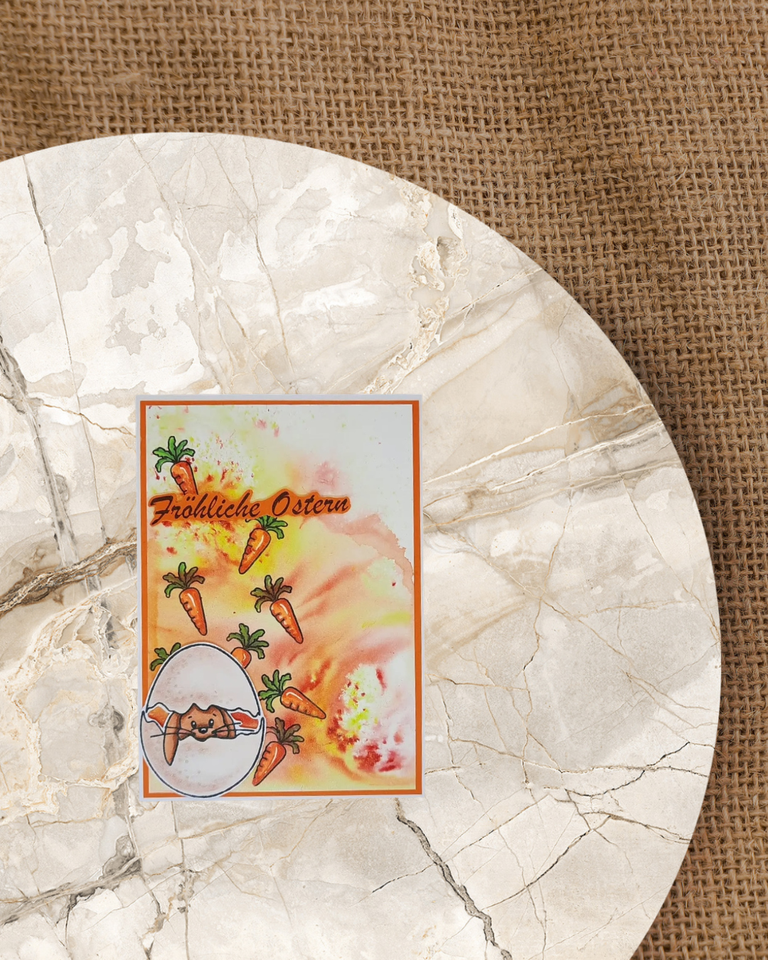 Karte mit einem Hasen der aus einem zerbrochenen Ei schaut. Im Hintergrund rötlich gelber Wasserfarbenhintergrund mit vielen Karotten. Schriftzug fröhliche Ostern