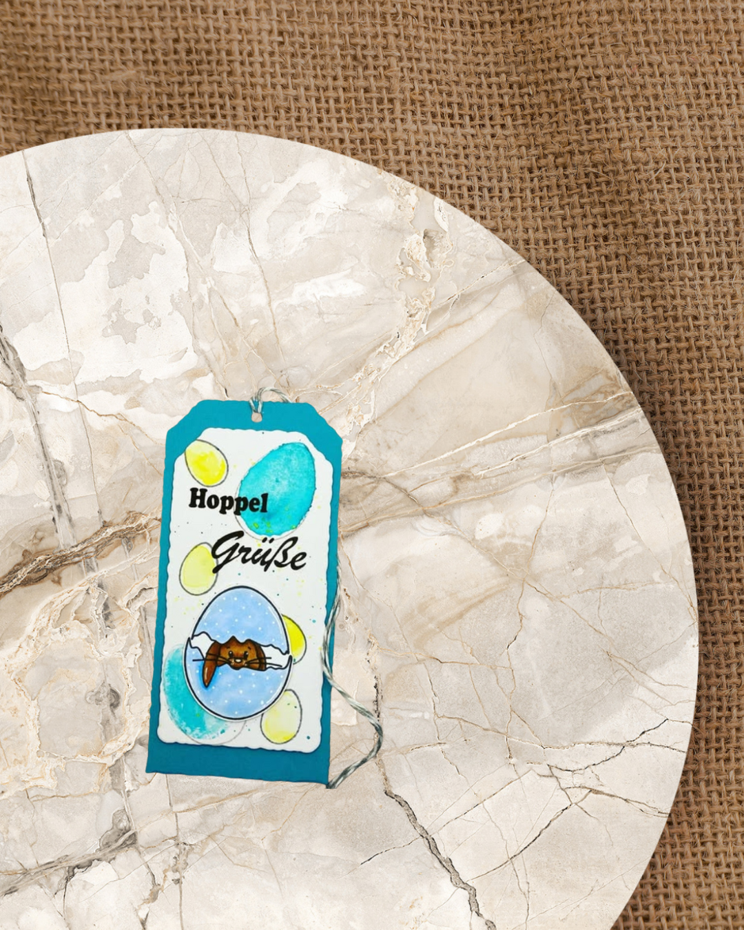 Oster Anhänger mit einem Hasen der aus einem zerbrochenen Ei schaut. Im Hintergrund verdienen große Eier in gelb und blau. Spruch Hoppel Grüße
