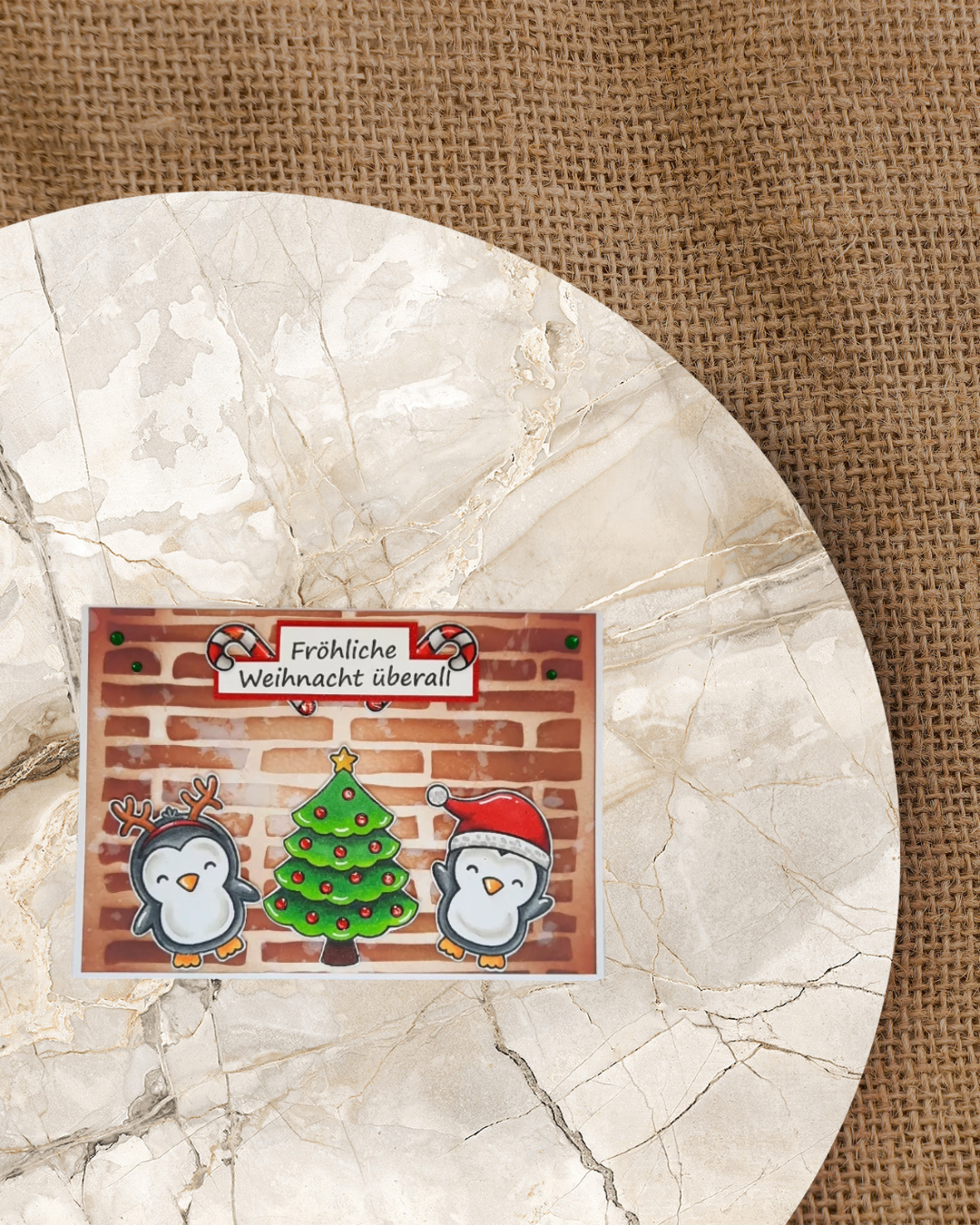 Karte mit 2 Pinguinen die ein Geweih und eine Mütze aufhaben. In der Mitte ein Christbaum mit Kugeln und Stern. Als Hintergrund ist eine braune Mauer. Spruch Fröhliche Weihnachten und jeweils eine Zuckerstange auf der Seite