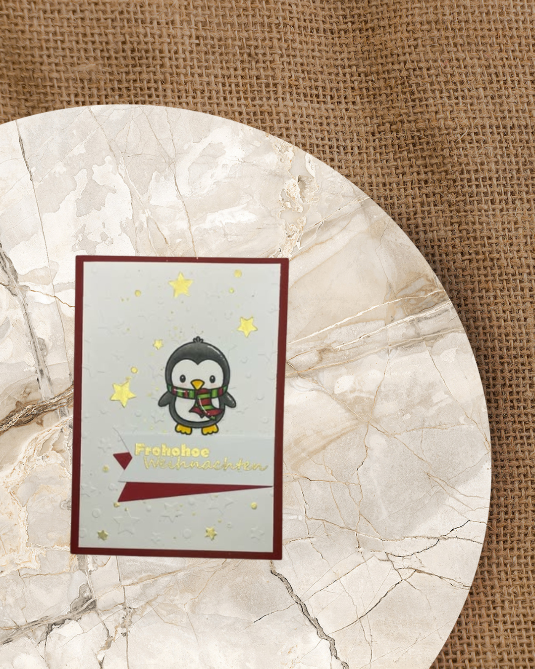 Pinguin mit Schal und Frohe Weihnachten. Im Hintergrund golden Sterne und Rot unterlegt.