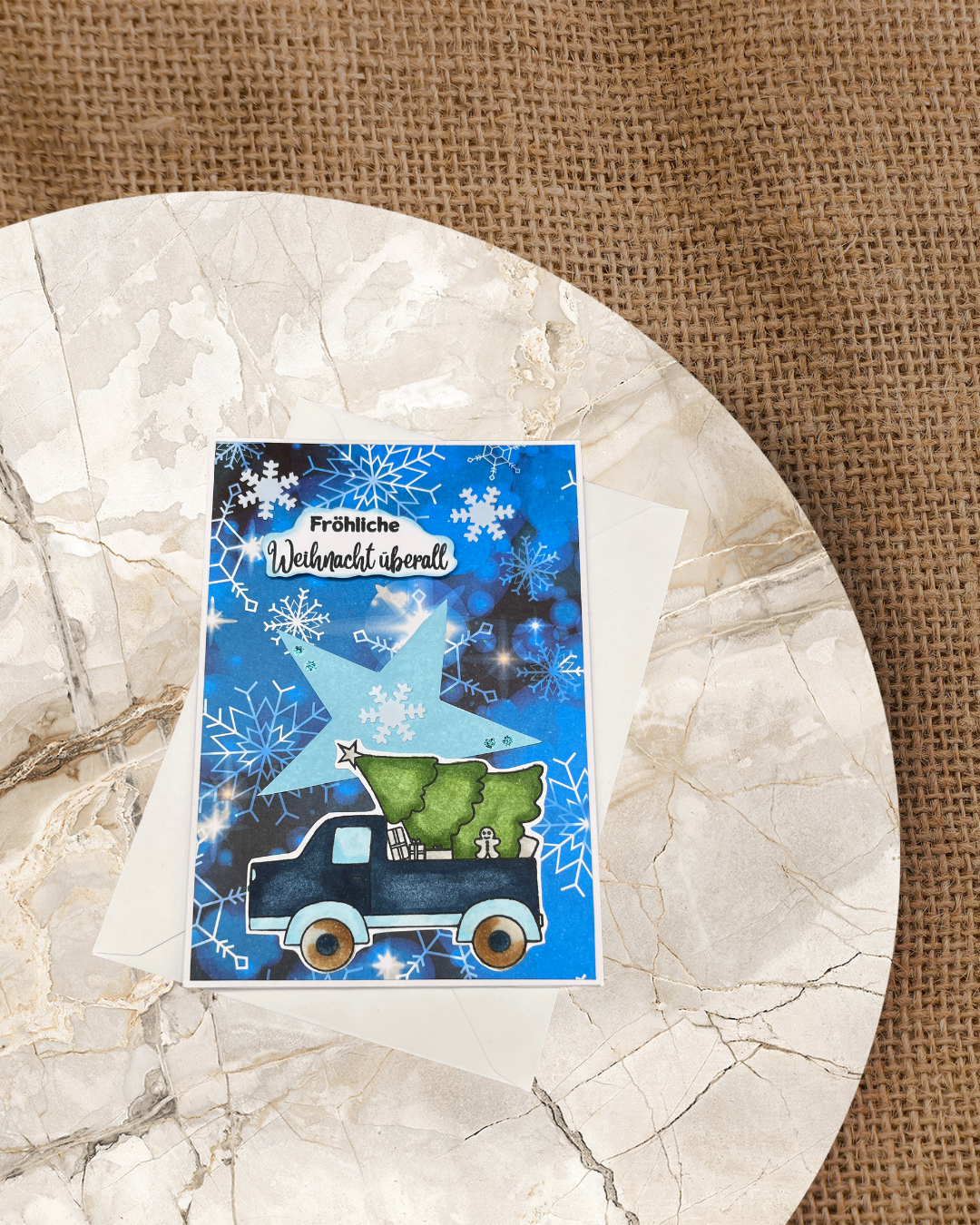 Karte in Blau einem Weihnachtstruck der einen Tannenbaum, Geschenke, Lebkuchenmann auf dem Truck hat. Im Hintergrund Sterne und Eiskristalle.