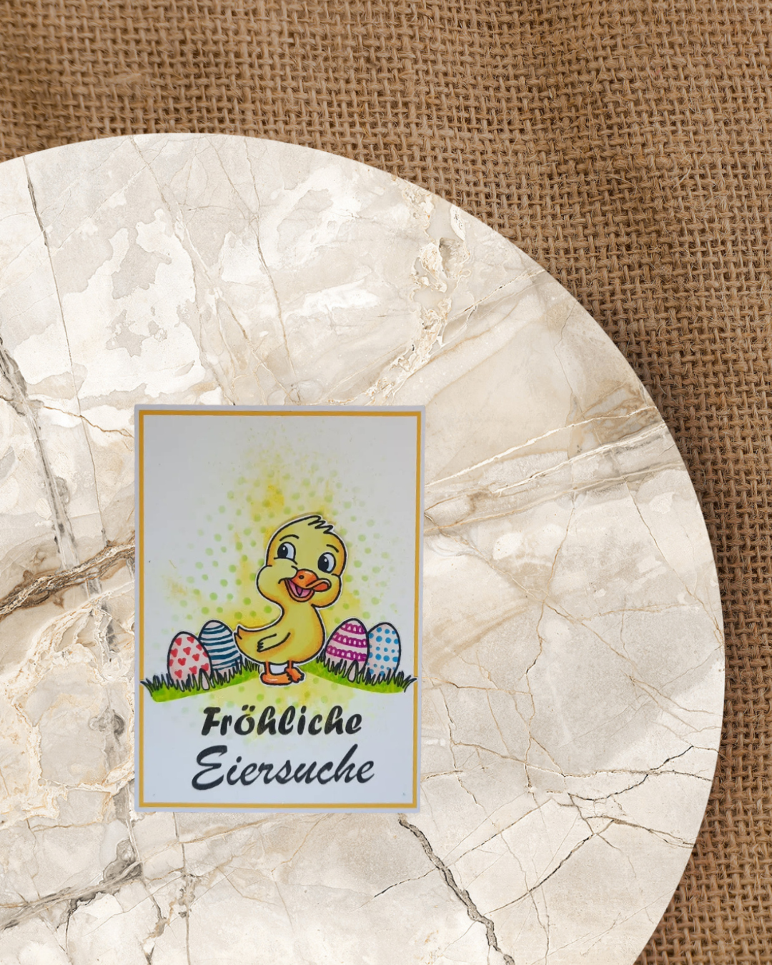 Entenküken in der Mitte auf einer Karte mit jeweils 2 bunten Eiern rechts und links mit Graß. Spruch Fröhliche Eiersuche