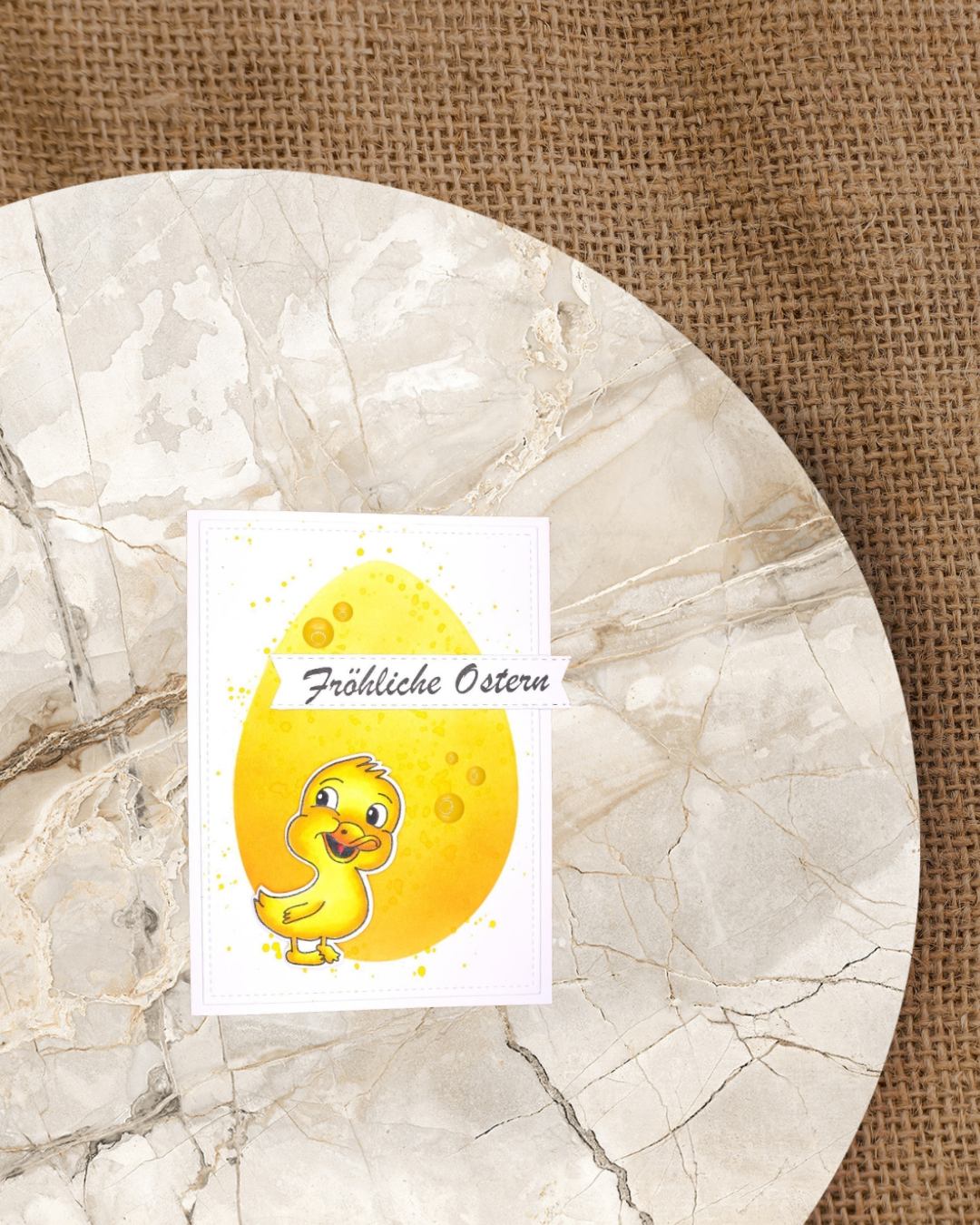 Auf einer Karte ein Entenküken mit einem großen gelben Ei als Hintergrund. Spruch Fröhliche Ostern