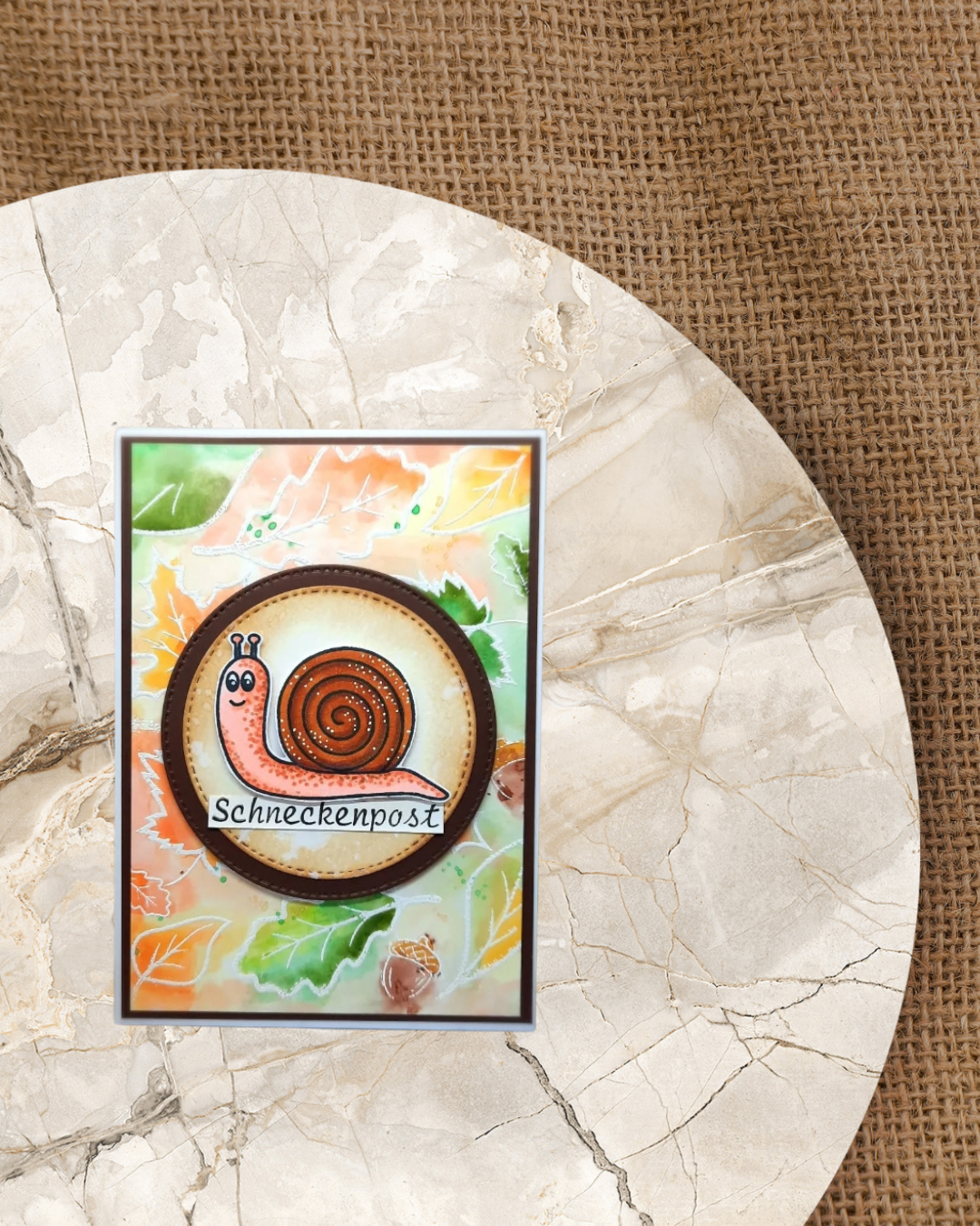 Karte mit einem Kreis, darin sitzt eine Schnecke. im Hintergrund ein herbstlich bunter Hintergrund mit Blättern. Unter de Schnecke steht Schneckenpost