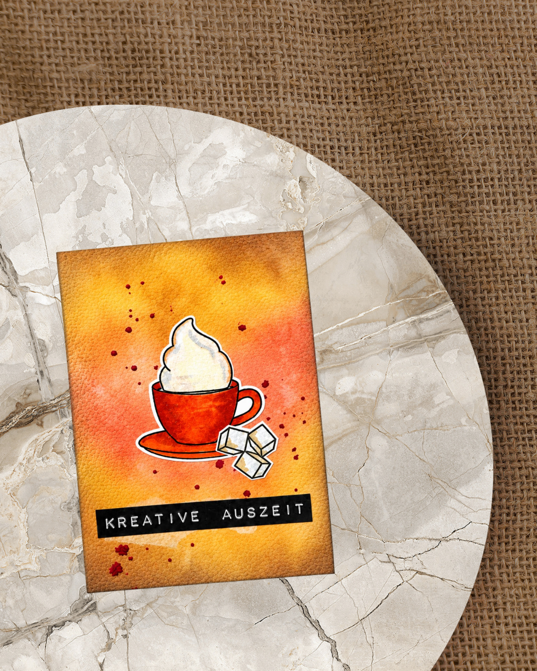 Handgemachte Karte mit gestempelter roter Kaffeetasse mit üppiger Sahnehaube und Zuckerwürfeln auf warmem Orange-Gelb-Distress Oxide-Hintergrund mit roten Farbspritzern und schwarzem Schriftzug „Kreative Auszeit" im unteren Bereich.