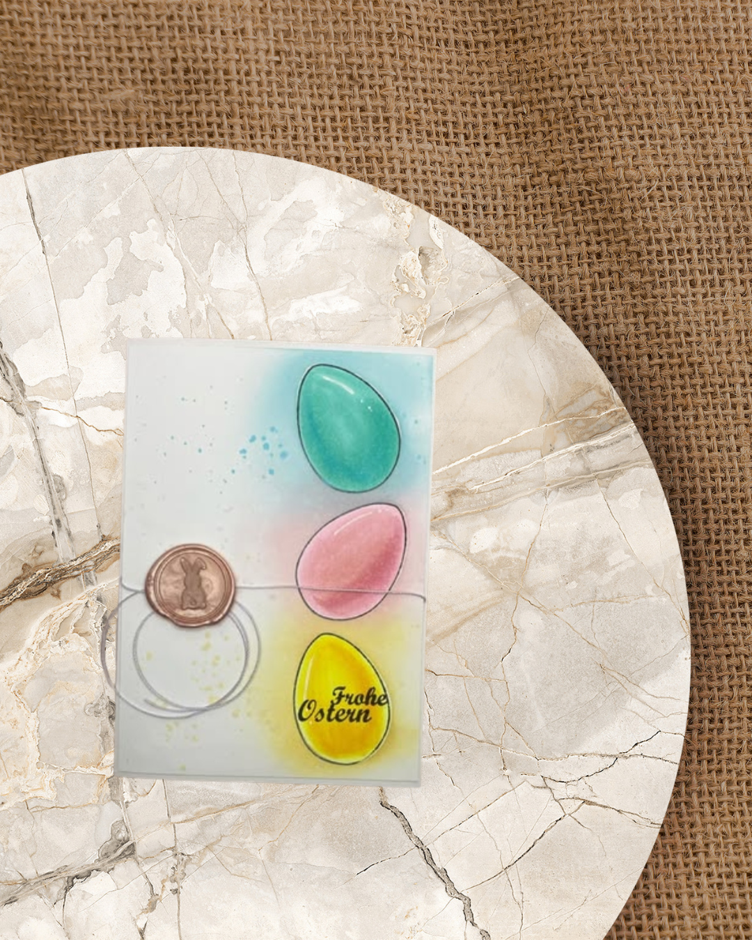 Karte mit 3 eiern aufeinander gestapelt in gelb, rosa, türkis. Siegelwachsstempel mit einen Hasen drauf und Spruch Frohe Ostern