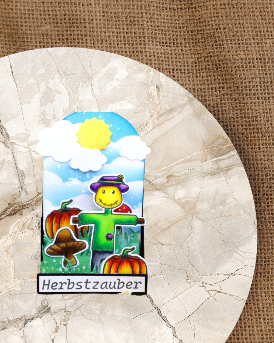 Anhänger mit einer Vogelscheuche, Pilzen, Kürbisse drauf. Im Hintergrund Wolken und eine Sonne. Spruch Herbstzauber