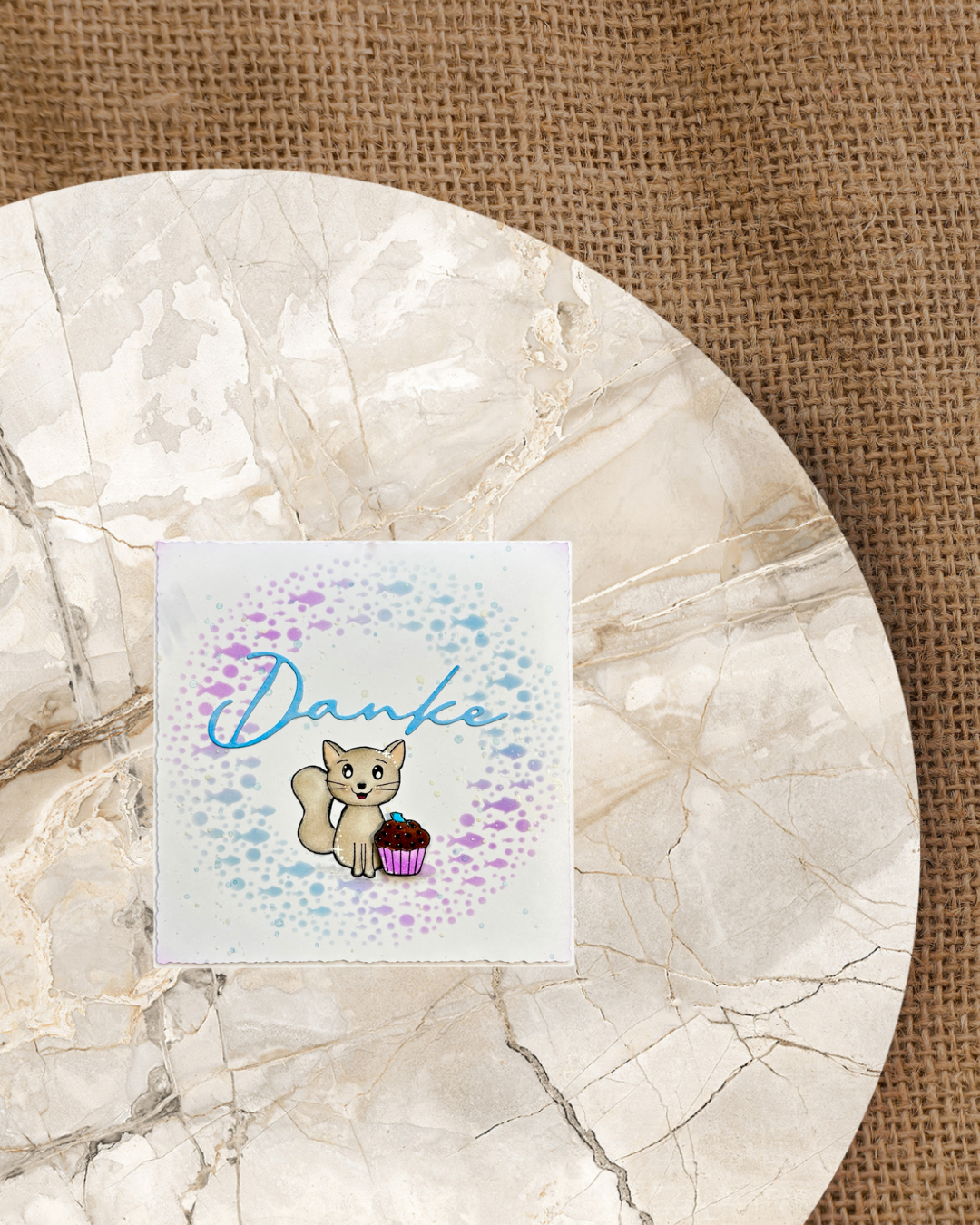 Sitzenden Katze mit Cupcake. Als Hintergrund ein Fischkreis mit Blau und lila mit großem Schriftzug Danke