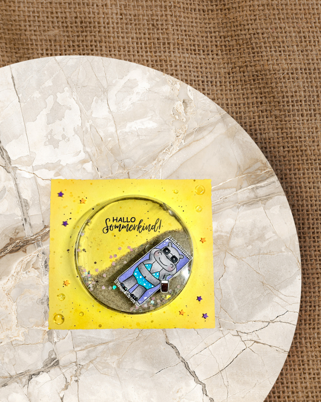 Gelbe Schüttelkarte. Das Schüttelelement ist mit Sand und Muscheln gefüllt. Darauf ist ein Hippo auf einen Strandtuch mit Bikini und Sonnenbrille. Spruch Hallo Sommerkind.