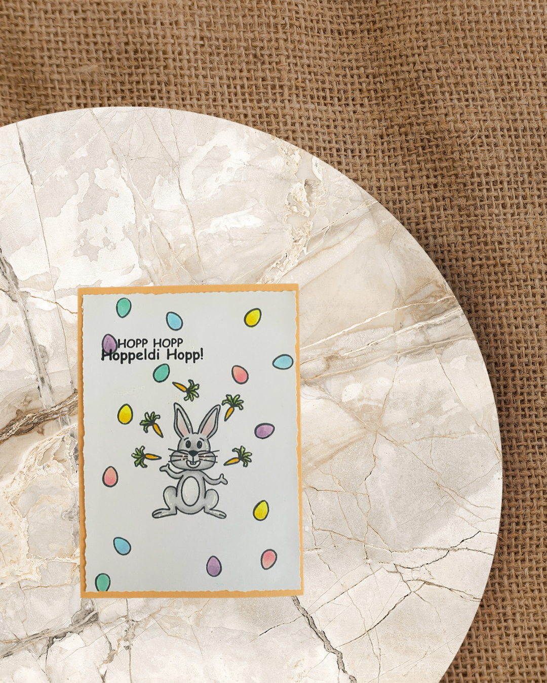 In der Mitte der Karte ein sitzender Hase der jongliert mit Karotte. Im Hintergrund viele bunte Eier. Spruch Hopp Hopp Hoppeldi Hopp