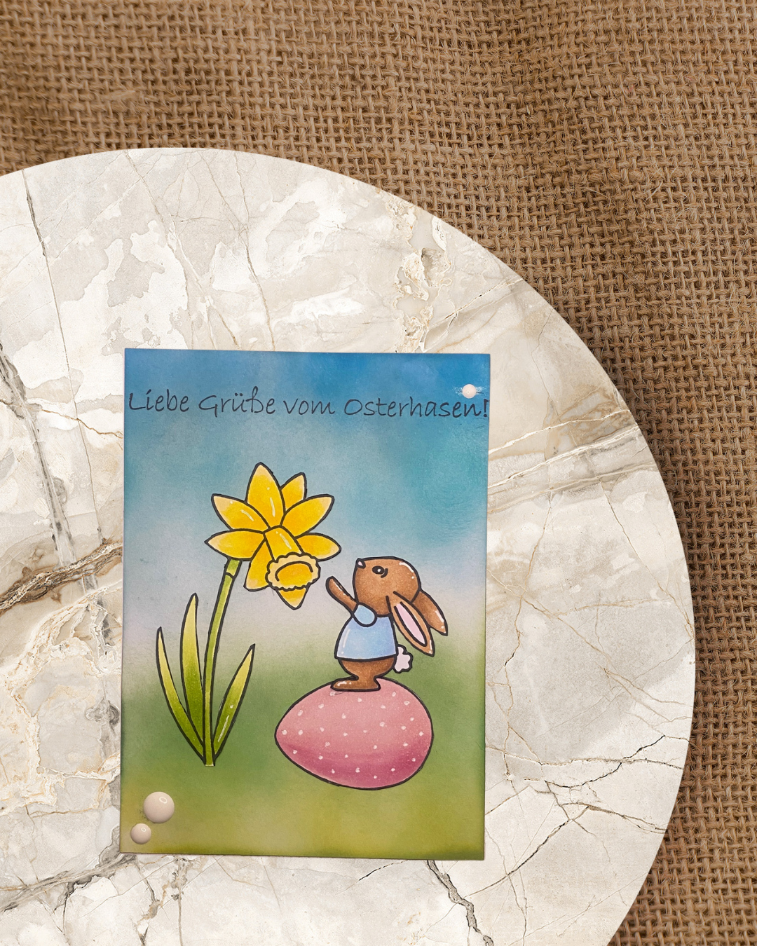 Karte mit Hase auf Ei stehend an einer Blume mit Text Liebe Grüße vom Osterhasen 