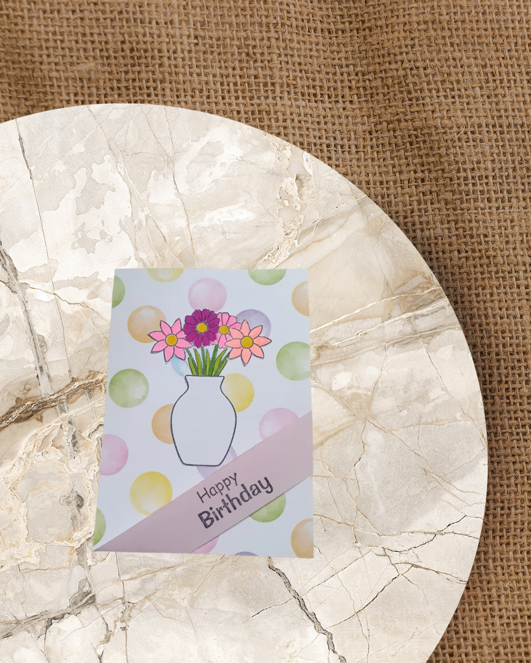 Karte mit bunten Punkten im Hintergrund. Im Vordergrund eine Vase mit einem Frühlingsblumenstrauß in den Farben Rosa und Lila. Über die rechten unter Kecke ein Band mit dem Spruch Happy Birthday.