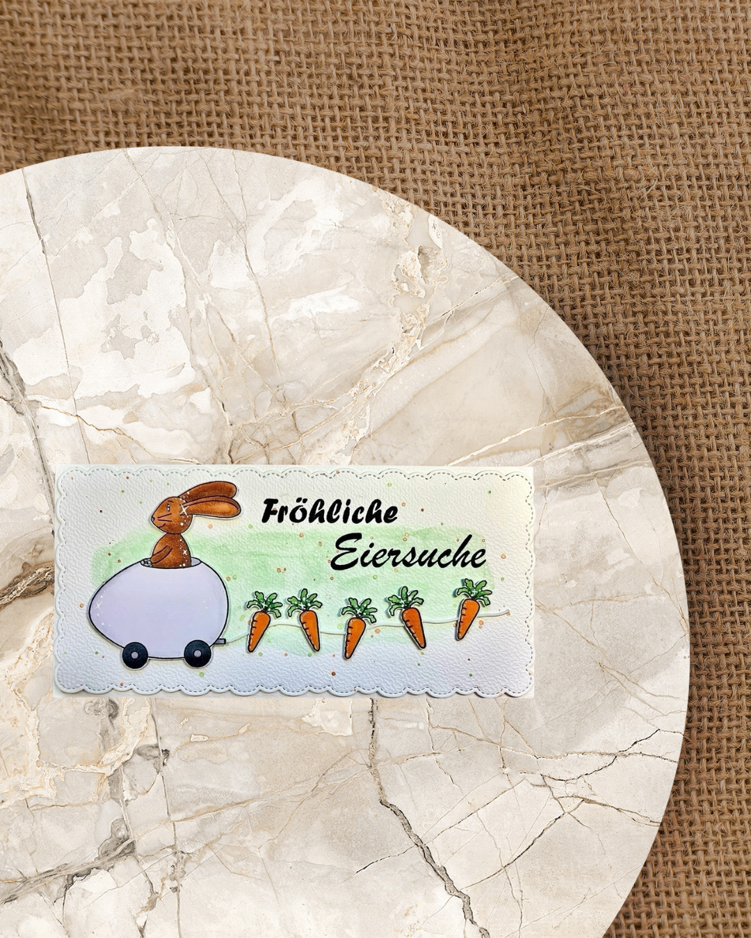 Karte für Ostern mit einem Hase der in einem Eiauto sitzt und eine Leine mit Karotten hinter sich her zieht. Spruch Fröhliche Eiersuche 