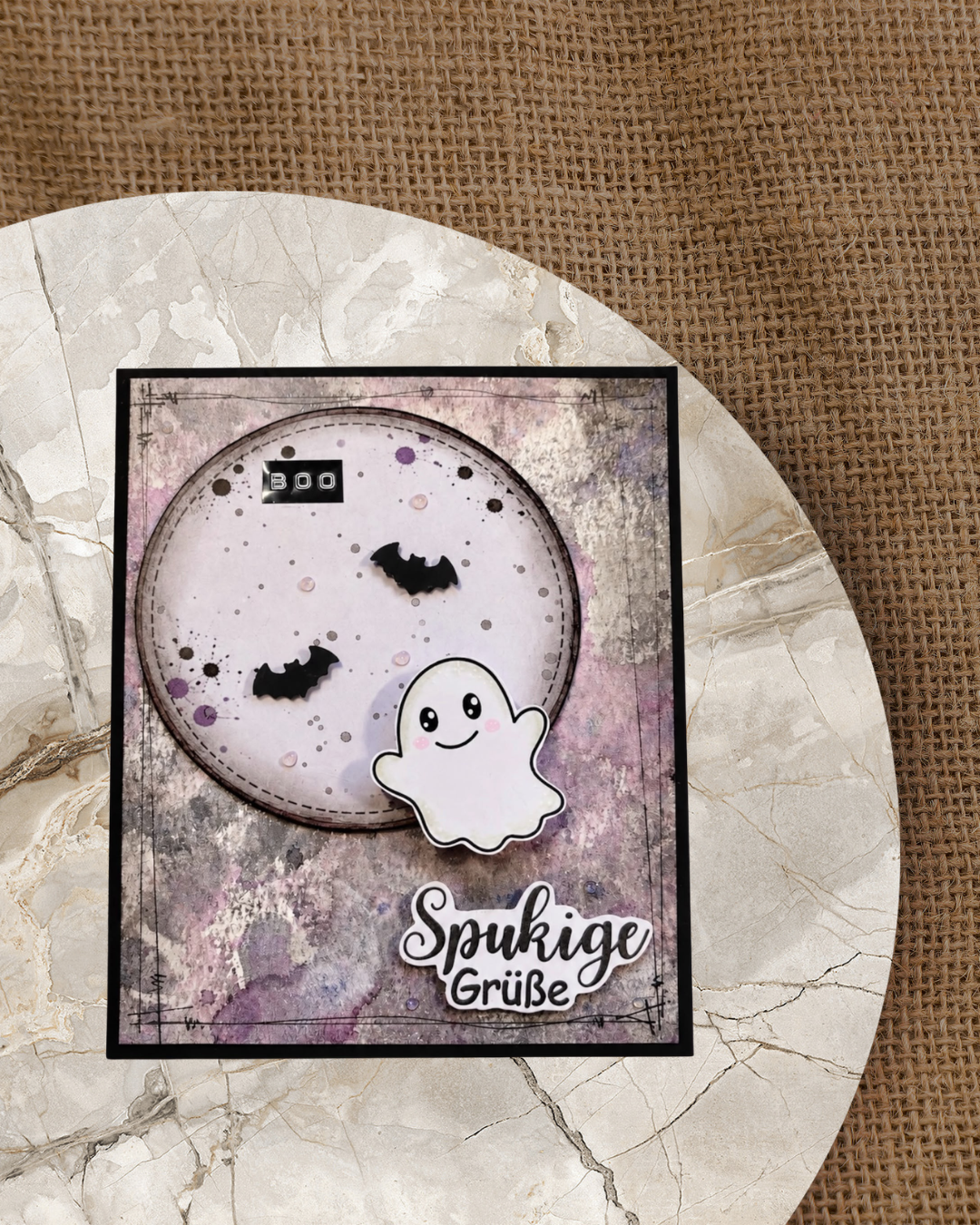 Handgemachte Halloween-Karte mit niedlichem weißen Geist und zwei schwarzen Fledermäusen auf rundem Mondausschnitt, eingebettet in einen lila-grauen Aquarell-Hintergrund mit „BOO"-Schriftzug und „Spukige Grüße" als ausgeschnittenen Schriftzug im Vordergrund.