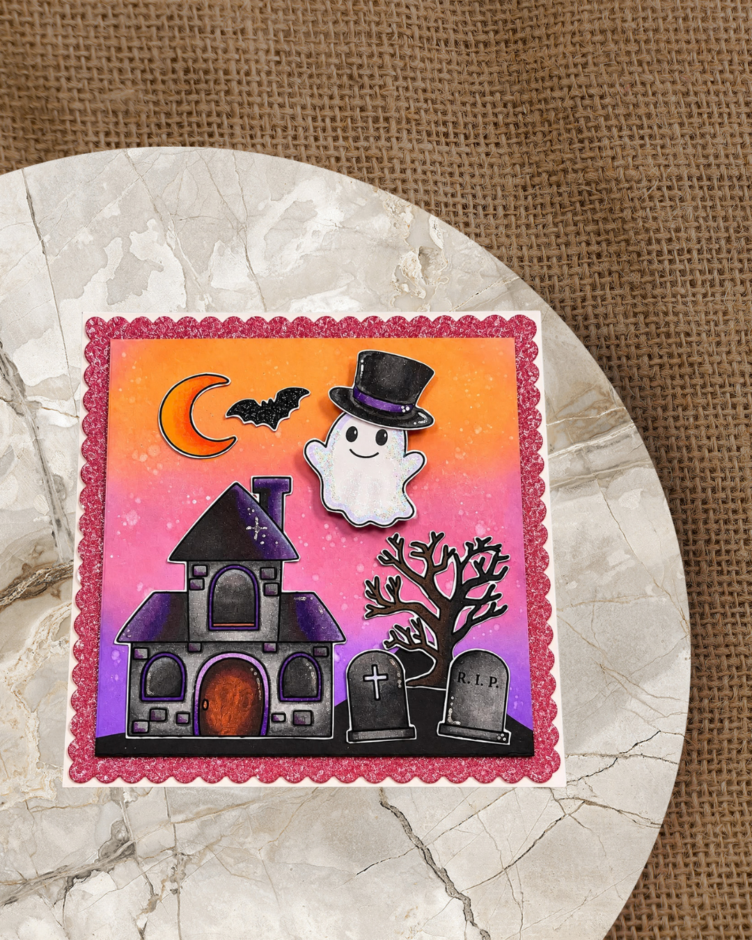 Handgemachte Halloween-Karte mit niedlichem Geist im schwarzen Zylinder, dunklem Geisterhaus, knorrigem Baum, zwei Grabsteinen mit Kreuz und R.I.P., Fledermaus und orangefarbenem Halbmond auf warmem Orange-Pink-Lila-Verlauf, eingerahmt von einem rosa Glitzerrand.