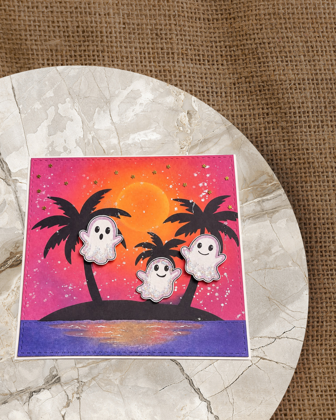 Handgemachte Halloween-Karte mit drei niedlichen Glitzer-Geistern vor einer tropischen Sonnenuntergangs-Silhouette mit schwarzen Palmen und großem orangefarbenem Mond auf leuchtendem Pink-Orange-Verlauf mit goldenen Sternen und lila Wasserreflexion.