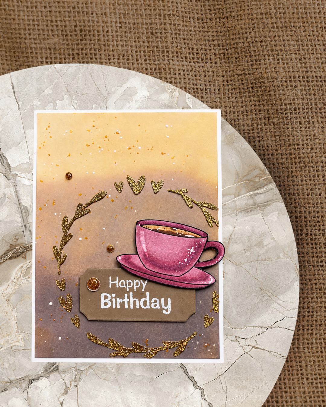 Handgemachte Geburtstagskarte mit gestempelter rosa Kaffeetasse auf Untertasse, umrahmt von goldenen Glitzerblättern und Herzen auf warmem Ombre-Hintergrund in Gelb und Lila, mit „Happy Birthday" Schriftzug auf braunem Etikett.