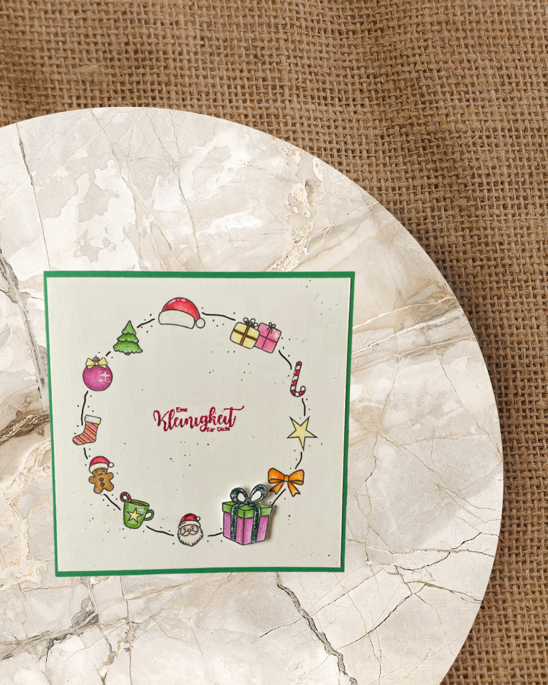 Karte mit einem Kreis gebildet mit verschiedenen Weihnachtsmotiven wie z.B. Nikolausmütze, Tannenbaum, Geschenke, Schleife, Christbaumkugel. In der Mitte vom Kreis ein Spruch in rot mit Eine Kleinigkeit für Dich. 